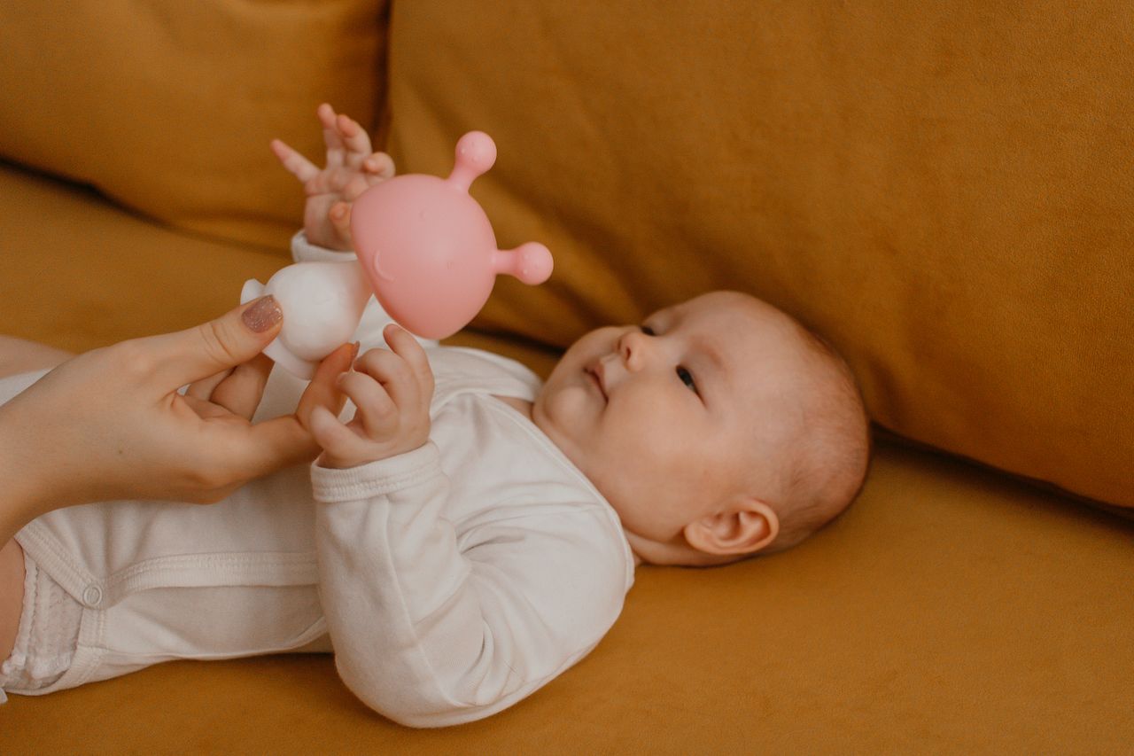 A baby lying on a couch playing with a pink and white toy, engaging with an adult hand.
