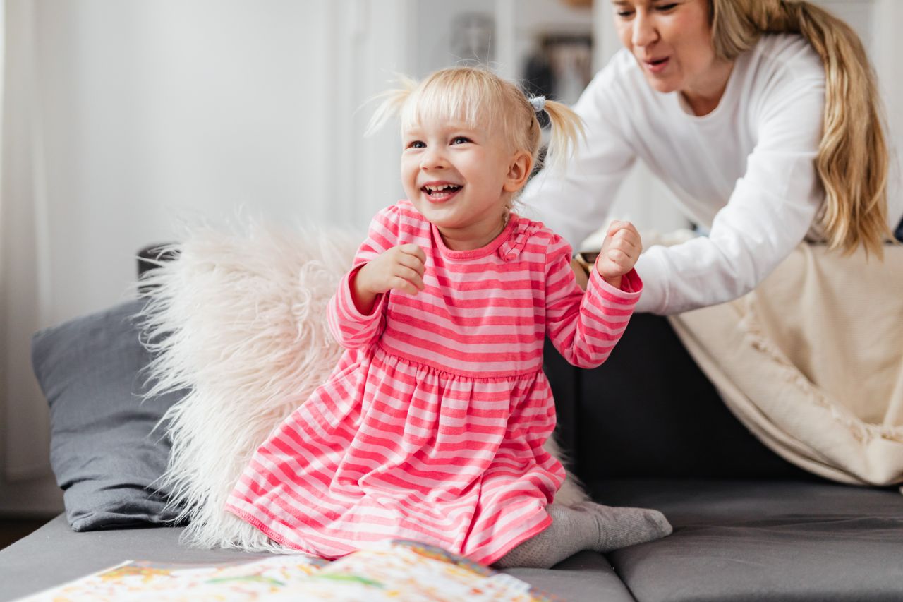 Joyful toddler girl in pink dress playing on a couch with her mother indoors.